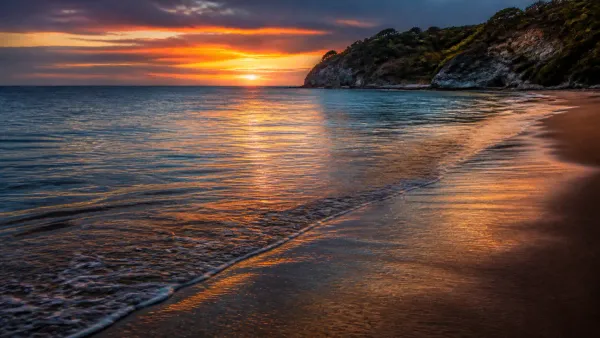 Peaceful shoreline at twilight with gentle rhythmic ripples lapping on dark wet sand reflecting glowing amber and gold evening sky