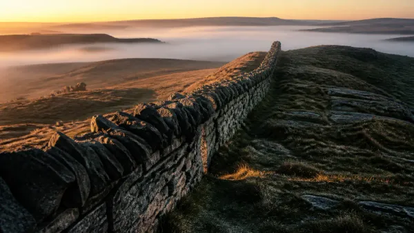 Long weathered dry-stone wall receding across open highland fells into morning mist as raking amber dawn light catches the stones